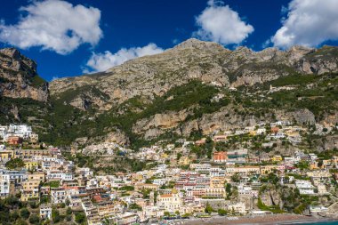 İtalya 'nın Positano kentindeki parlak Akdeniz binaları dramatik kireçtaşı dağlarının altındaki dik yamaçlar boyunca istiflenmiştir. Sahnede canlı yüzler, yemyeşil yeşillikler ve Amalfi Sahili 'nin üzerinde dağınık bulutlar olan koyu mavi bir gökyüzü yer alıyor..