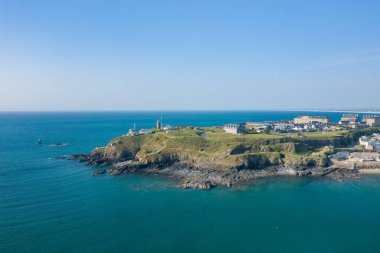 Pointe du Rocs 'un hava manzarası engebeli bir deniz feneri, çimenli yamaçlar ve berrak bir gökyüzünün altında canlı mavi denizle çevrili kayalık bir kıyı şeridi. Sahne açık, güneşli ve geniş kıyı dokularıyla huzurlu..