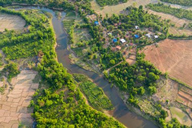Tepedeki görüntü kıvrımlı bir nehir, yeşil orman arazileri ve ekili tarlalarla çevrili küçük bir köy gösteriyor. Manzara bereketli bitki örtüsü, geometrik tarım arazisi ve yumuşak gün ışığı altında sakin, doğal bir atmosfere sahiptir..