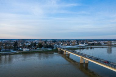 Çağdaş bir köprü geniş Loire Nehri boyunca uzanır ve yumuşak mavi bir gökyüzünün altında karla kaplı çatıları ve ağaçları olan bir köye götürür. Sakin su, Sully sur Loire 'ın sakin kış atmosferini yansıtıyor..
