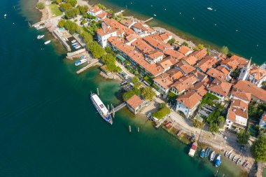 Isola dei Pescatori 'nin hava görüntüsü bir terracotta çatısı kümesi, bir kilise kulesi ve teknelerle kaplı yoğun bir rıhtım olduğunu gösteriyor. Ada, Maggiore Gölü 'nün berrak mavi sularıyla çevrili, yemyeşil bir zemin üzerine kurulmuş.