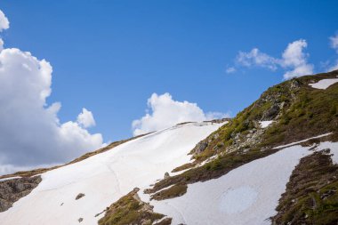 Aiguillette des Houches yamaçlarında eriyen karlar, açıkta kalmış kayalar ve dalgalı kümülüs bulutları ile mavi gökyüzünün altında yeşil bitki örtüleri bulunur..