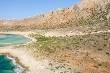 Yunanistan 'ın Girit kentindeki Balos Sahili' nin turkuaz sularının üzerinde seyrek yeşil bitki örtüsü yükselen güneşli kayalık tepeler. Manzara kurak, dokulu ve açık mavi bir gökyüzü altında genişliyor..