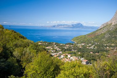 Berrak bir gökyüzünün altında engebeli dağları olan, derin mavi Tyrhenian Denizi 'ne bakan yemyeşil tepelerin arasında yuva yapmış bir sahil köyünün geniş manzarası. Parlak güneş ışığı canlı renkleri ve sakin Akdeniz atmosferini güçlendirir.