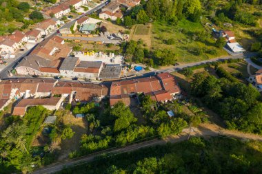 Yeşil bitki örtüsü ve açık arazilerle çevrili Saint-Maurice-sous-les-Cotes kırsalındaki geleneksel kırmızı kiremitli çatıların ve küçük bahçelerin hava manzarası. Kompozisyonda evlerin yama düzeni, yaz yaprakları ve bir