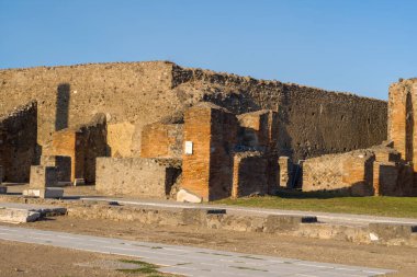 Altın öğleden sonra ışığı, İtalya 'nın Pompeii kentindeki arkeolojik kamaralarda yıpranmış tuğla ve volkanik taş duvarları aydınlatıyor. Tarihi yapılar, açık mavi gökyüzünün altında desenli masonluk ve geometrik desenleri ortaya çıkarıyor..
