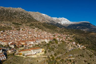 Yunanistan 'ın Arachova kentinin hava manzarası, engebeli bir yamaç boyunca yayılmış yoğun bir kırmızı çatılı ev kümesini, etkileyici kar örtülü dağların parlak mavi gökyüzünün altında yükseldiğini gösteriyor..