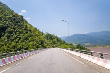 Geniş asfalt köprüsü, yemyeşil tepeleri olan çamurlu bir nehri ve Kuzey Vietnam 'da açık bir gökyüzünün altındaki uzak mavi dağları geçiyor. Parlak güneş ışığı canlı renkleri ve açık, huzurlu atmosferi güçlendirir..