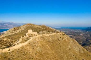 Yunanistan 'da berrak bir gökyüzü altında engin bir kırsal alan ve uzak mavi denize bakan, Akrokorint' in altın yamaçlarını kaplayan yıpranmış kale harabeleri ve taş duvarlardan oluşan havadan bir perspektif..
