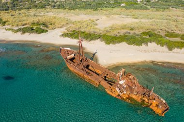 Yunanistan 'ın Gythio kenti yakınlarındaki kumlu bir sahil şeridi boyunca sığ turkuaz sularda sıkışmış paslanmış Dimitrios gemi enkazının belirsiz hava manzarası. Terk edilmiş gemi, parlak güneş ışığı altında canlı deniz ve doğal yeşillikle çelişiyor..