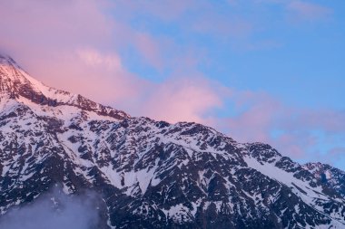 Şafak vakti Aiguille de Bionnassay 'in engebeli, karlı yamaçlarında, yumuşak pembe bulutlar süzülür, dramatik alp manzarasını aydınlatan hafif ışıkla..