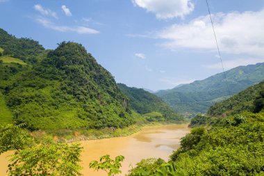 Kıvrımlı sarı bir nehir yemyeşil tepeler ve Kuzey Vietnam 'daki uzak yeşil dağlar arasında akar. Parlak güneş ışığı ve dağınık bulutlar zengin dokular ve doğal karşıtlıklarla dolu canlı, yaz manzarası yaratır..
