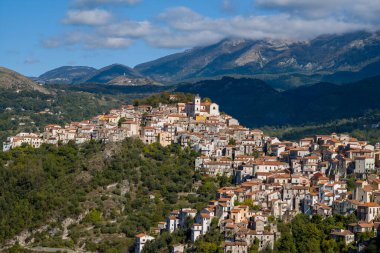 İtalya 'nın Rivello kenti, yoğun kümelenmiş taş evler ve terasotta çatılarıyla yemyeşil bir yamacın tepesinde dağınık bulutlu mavi bir gökyüzünün altındaki engebeli dağların üzerine kurulmuştur..