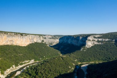 Gorges de lArdeche 'nin geniş hava manzarası dramatik dikey kireç taşı uçurumlarını, yoğun yeşil ormanı ve açık mavi gökyüzünün altında dolambaçlı bir nehri gösteriyor. Güneş ışığı engebeli dokulara ve geniş doğal manzaraya vurgu yapıyor..