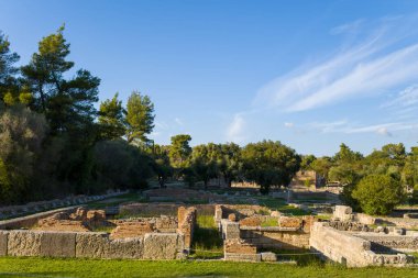 Yunanistan 'ın Olympia kentindeki yemyeşil ve olgun ağaçların arasında Leonidaion misafirhanesinin güneş taşı ve tuğla kalıntıları duruyor. Tarihi arkeolojik manzara, açık mavi bir gökyüzünün altında sıcak öğleden sonra ışığında yıkanır..