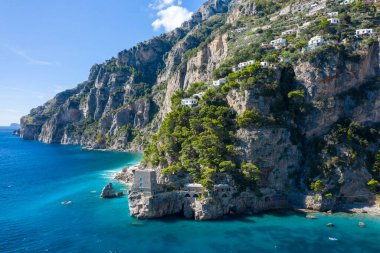 İtalya 'nın Positano kenti yakınlarındaki Amalfi Sahili' nin turkuaz sularının üzerinde yükselen dik kireçtaşı kayalıkları, verimli yeşilliklerin arasına yerleşmiş dağınık villaları barındırıyor. Sahnede engebeli kaya oluşumları, canlı Akdeniz yaprakları ve açık mavi bir deniz yer alıyor.