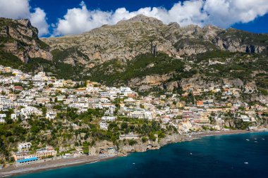 Amalfi kıyısındaki Positano, İtalya 'nın renkli teraslı evlerinin arkasında dik kireçtaşı dağları dramatik bir şekilde yükseliyor. Canlı yamaçlar, parlak bulutlarla benekli gökyüzünün altındaki derin mavi Akdeniz sularına bakıyor..