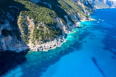 Yoğun yeşil bitki örtüsüyle kaplanmış dik kireçtaşı kayalıkları Akdeniz 'in Cala Goloritze, Baunei, Sardinya yakınlarındaki berrak, canlı mavi sularıyla buluşmak için iniyor. Güneşli sahil şeridi kayalık dokular ve...
