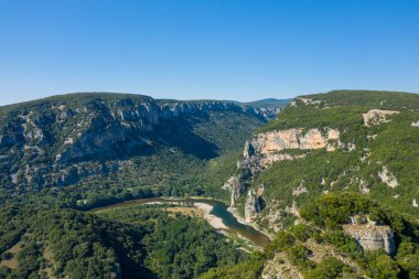 Gorges de Ardeche 'deki dolambaçlı bir nehrin üzerinde yükselen dramatik kireçtaşı uçurumlarının havadan görünüşü. Yoğun yeşil orman, berrak mavi bir gökyüzünün altındaki yamaçları kaplıyor ve canlı bir yaz manzarası yaratıyor..