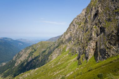 Dik kayalık uçurumlar Pireneler 'deki güneşli yeşil bir tepenin üzerinde yükseliyor, uzak sarmal vadiler ve berrak mavi bir gökyüzü engin ve doğal bir güzellik hissi yaratıyor..