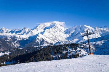 Güneşli bir yamaçtan yükselen bir kayak asansörü, Mont Blanc kitlesinin panoramik manzarasına ve canlı mavi gökyüzünün altındaki derin yeşil ormanlara sahiptir. Kış ışığı dramatik alp manzarasını ve el değmemiş karı güçlendirir..