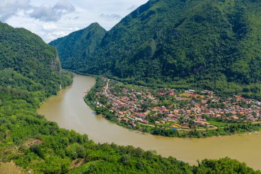 Laos 'un Nong Khiaw kentinin geniş bir hava manzarası Nam Ou nehrinin sarp yeşil kireçtaşı dağları ve yoğun tropikal ormanlarla çevrili canlı bir köyde kıvrıldığını gösteriyor. Sahnede geleneksel çatılar, yemyeşil bitki örtüsü ve dramatik