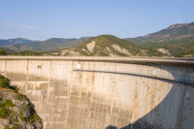 Barrage de Castillon 'un geniş manzarası geometrik gölgeli beton duvar, güneşli yeşil tepeler ve engebeli dağların altında berrak bir gökyüzü..