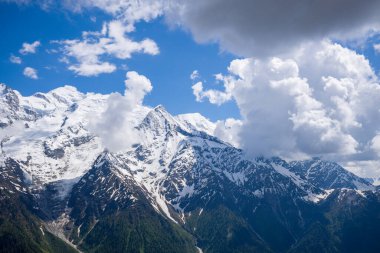 Impressive cumulus clouds cast dramatic shadows over the snow-covered slopes and rugged ridges of the Mont Blanc massif, with patches of green forest visible below under vibrant blue sky.