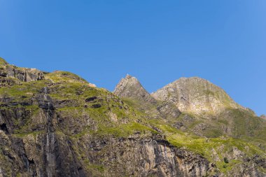 Güneşli, çimenli yamaçların ve Gavarnie Sirki 'ndeki engebeli kayalıkların üzerinde berrak mavi bir gökyüzüne karşı uzanan pürüzsüz bir tepe yükselir. Sahnede doğal dokular, canlı renkler ve çarpıcı yükseklik yer alıyor..