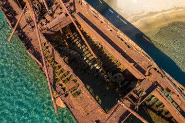 Dimitrios 'un enkazının kumlu bir Yunan kıyı şeridi yakınlarındaki şeffaf turkuaz suyla paslanmış iskelet kalıntılarının genel manzarası. Güneş ışığı, aşınmış kirişleri, açık gövde yapısını ve gölge ile ışığın etkileşimini vurgular..