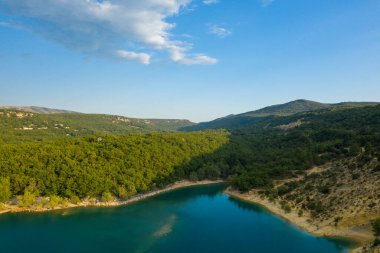 Sainte-Croix gölünde berrak bir gökyüzünün altında berrak mavi sularla çevrili sakin bir körfez, yoğun yeşil ormanlarla ve yumuşak tepelerle çevrilidir..