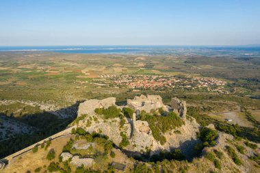 Güneşli bir tepenin tepesindeki yıpranmış taş kalıntılarının havadan görünüşü yamalı tarlalara, bir köye ve Opoul Perillos yakınlarındaki uzak mavi denize bakıyor..