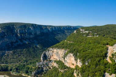 Engebeli kireçtaşı uçurumlarının ve Gorges de Ardeche 'deki bir platodaki yoğun yeşil ormanın havadan görünüşü. Parlak güneş ışığı keskin gölgeler bırakır dramatik araziyi ve açık mavi gökyüzünü aydınlatır..