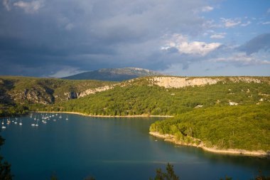 Karamsar gökyüzü dramatik bulutlarla aydınlanıyor sakin mavi sular boyunca kayıyor ve Sainte-Croix Gölü 'nün ormanlık kıyı şeridinde demirli yelkenlileri ve engebeli uçurumları var..