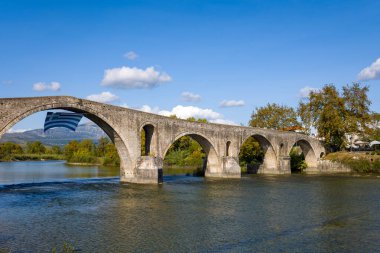Antik kemerli taş köprü Yunanistan 'ın Arta kentinde sakin bir nehir boyunca uzanırken, kemerlerinden birinin altında bir Yunan bayrağı asılı bulunuyor. Sahnede açık mavi gökyüzü, dağınık bulutlar ve yumuşak gün ışığında yemyeşil nehir kenarındaki ağaçlar yer alıyor..