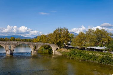 Tarihsel çok kemerli taş köprü, Yunanistan 'ın Arta kentinde nehir kenarındaki kafeleri altın sonbahar ağaçları altında büyük şemsiyelerle gölgelendirilmiş temiz bir nehir boyunca uzanır. Uzak dağlar ve dağınık bulutlu mavi bir gökyüzü sakin manzarayı yumuşak bir şekilde tamamlıyor.