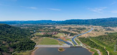 Geniş hava manzarası, açık ovalar, ormanlık tepeler ve açık mavi gökyüzünün altındaki uzak dağ sırtlarıyla çevrili kırmızı çatılı binaların yanında bir baraj ve kanal sistemi içerir. Manzara kesişme noktasını vurgular
