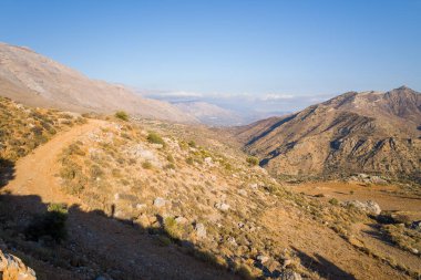Yunanistan 'ın Girit kentindeki engebeli bir dağ yamacı boyunca açık mavi gökyüzü altında kuru kayalık yol kıvrımları. Sıcak güneş ışığı kurak dokulara ve uzak tepelere vurgu yaparak açıklık ve macera hissi yaratıyor..