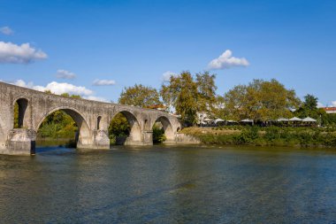 Tarihsel çok kemerli taş köprü Yunanistan 'ın Arta kentinde sakin bir nehir boyunca uzanırken, nehir kenarındaki yapraklı ağaçlar ve berrak mavi bir gökyüzü sakin bir sonbahar atmosferi yaratıyor. Sahne, yumuşak gün ışığında dokulu taş işçiliğini ve sakin suyu ön plana çıkarıyor..