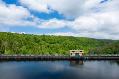 Sarı bir kamu binası, durgun ve karanlık sulara bakan beton bir barajın üzerinde duruyor. Yoğun yeşil ormanlar ve Chaumecon gölünde dağınık bulutlarla dolu bir gökyüzü..
