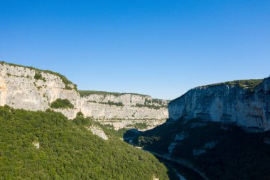 Dik kireçtaşı duvarlar dolambaçlı bir nehrin ve Ardeche Boğazı 'nda yoğun bir ormanın üzerinde bulutsuz mavi bir gökyüzünün altında yükselir. Parlak güneş ışığı engebeli kaya dokusunu ve derin gölgeleri vurgular..