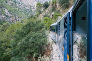 Yunanistan 'ın Peloponnese bölgesinde yoğun yeşil ağaçlar ve engebeli yamaçlarla çevrili kayalık bir dağ yolu boyunca mavi dar bir tren kıvrımları bulunuyor. Sahne, doğal tepe arazisi ve yemyeşil yapraklar boyunca uzanan dramatik demiryolu rotasını vurguluyor..