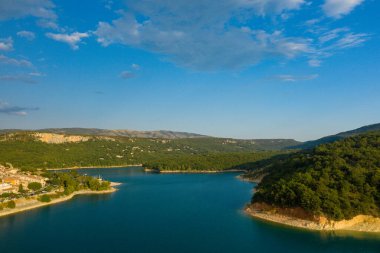 Bereketli yeşil tepeler ve Bauduen yakınlarındaki kayalık uçurumlarla çevrili derin mavi sularla dolu sakin bir körfezin geniş hava manzarası. Dağınık bulutlarla dolu temiz bir gökyüzünün altında..