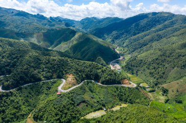 Laos 'taki Phou Khoun ve Luang Prabang arasında yeşil tepeler ve vadilerden geçen kıvrımlı dağ yolu yılanları. Yoğun orman, katmanlı tepeler ve dağınık kırsal evler dağınık bulutlarla dolu canlı mavi bir gökyüzünün altında görülebilir..