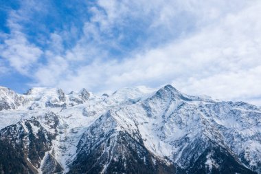 Broad view of the Mont Blanc massif with dramatic snow-laden ridges and forested lower slopes beneath a sky filled with layered, wispy clouds.