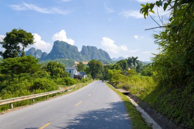 A quiet asphalt road curves through lush green countryside with dramatic limestone peaks in the distance near Mai Chau, Vietnam. Bright sunlight and blue sky enhance the tropical foliage and tranquil rural atmosphere.