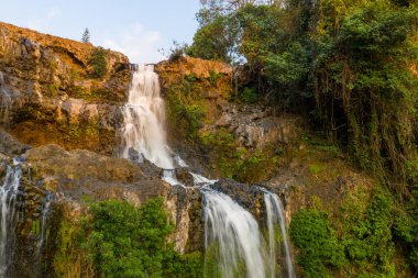 Sıcak akşam ışığı, Güney Laos 'taki Bolaven Platosu' nda yoğun yeşil yapraklarla çevrili engebeli bazalt uçurumlarından akan Tad Yuang şelalesi 'nin çağlayan sularını vurguluyor. Kaya yüzeyi ve canlı bitki örtüsü...
