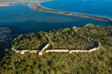 Yunanistan 'ın Navarino kenti yakınlarındaki sakin mavi bir lagünün üzerindeki yeşil, engebeli bir tepeyi çevreleyen antik taş kale duvarlarının insansız hava aracı görüntüsü, güneş ışığının manzarada uzun gölgeler oluşturması.