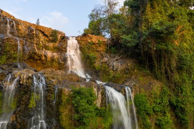 Sıcak akşam ışığı Tad Yuang şelalesinin çağlayan sularını, güney Laos 'taki Bolaven Platosu' ndaki yoğun tropik bitki örtüsüyle çevrili engebeli bazalt uçurumlarının üzerinden akarken yıkıyor. Kaya yüzeyi ve gür yeşillik çarpıcı bir etki yaratır.