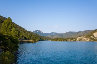 Bereketli yeşil tepelerle çevrili berrak mavi sularla dolu Castillon Gölü 'nün geniş manzarası ve parlak gökyüzünün altındaki engebeli tepeler..